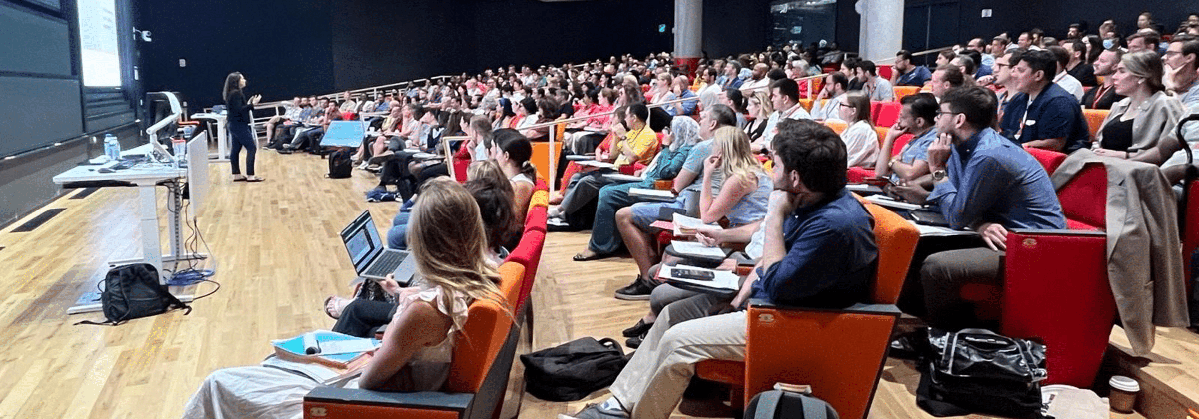 A large audience seated in a tiered auditorium during a lecture.
